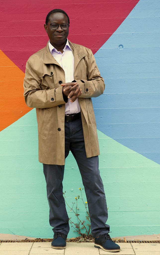 A man with dark skin standing in front of a brightly colored, geometric mural. He is wearing glasses, a light brown trench coat, a pale pink button-up shirt, and dark pants with black shoes. The individual has a calm expression, hands clasped in front, and the colorful mural behind them features vibrant shades of blue, red, orange, and green, creating a bold backdrop.
