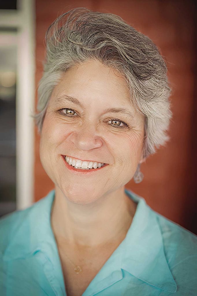 Headshot of Lisa Mount, a light-skinned woman with short gray hair, smiles warmly at the camera. She is shown from the chest up, wearing a light blue blouse, and her expression exudes friendliness and approachability. The background is softly blurred, featuring warm tones, which keeps the focus on her face. The overall composition suggests a candid and welcoming portrait.