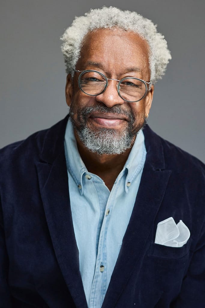 Headshot of Harry McCord, a brown-skinned man with gray curly hair and a beard is wearing round glasses and a friendly smile. He is shown from the chest up, dressed in a light blue denim shirt under a dark blue velvet blazer, with a white pocket square neatly folded in his jacket pocket. The background is a neutral gray, which draws attention to his warm expression and distinguished appearance. The image is a well-composed portrait that highlights his personality and style.