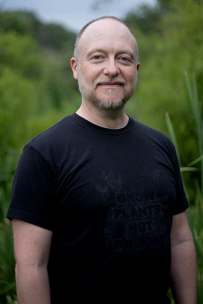 Headshot of Brooks Emanuel, a light-skinned man with a trimmed beard and short, thinning hair. He is shown from the waist up, wearing a black shirt and smiling slightly, with a blurred natural background of greenery. The soft lighting adds a calm and approachable feel to the portrait.