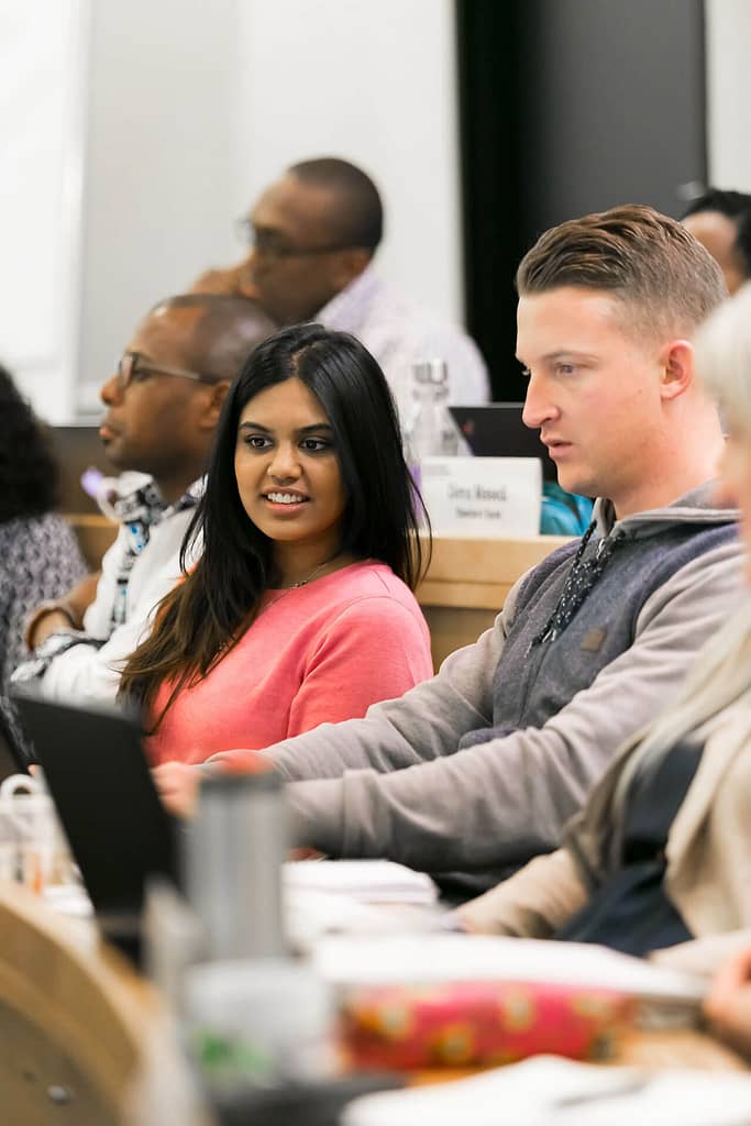 A group of people seated in a classroom or seminar setting. In the foreground, a woman with medium skin tone and long dark hair wears a bright pink top, sitting next to a man with light skin and short blonde hair, who is looking at a laptop. Behind them, other participants are engaged, including a man with dark skin and glasses in the background. The group appears to be focused on a presentation or discussion.