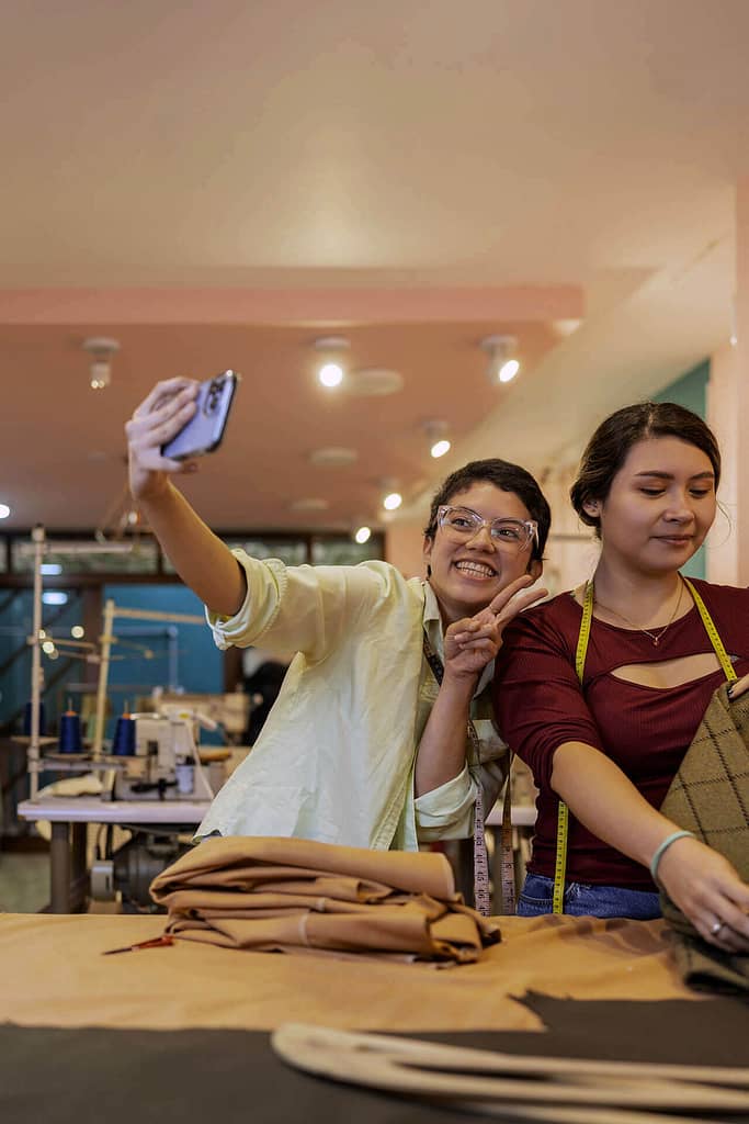 This image shows two young women in what appears to be a sewing or fashion workshop. The woman on the left, who has light brown skin and short dark hair, is smiling and holding up a phone to take a selfie, flashing a peace sign. They are wearing glasses and a light-colored shirt. The woman on the right, who also has light brown skin, dark hair tied back, and is focused on working with fabric, wears a measuring tape around their neck and a burgundy top. The background features sewing machines and folded fabric, giving the image a creative, workshop atmosphere.