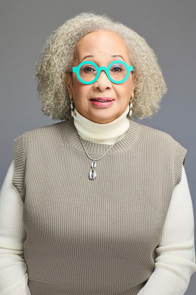 Headshot of Keryl McCord, a brown-skinned woman with gray curly hair wearing bright turquoise glasses, a beige sleeveless sweater over a cream turtleneck, and a silver necklace with a matching pendant. She is shown from the chest up, smiling slightly and looking directly at the camera, with a neutral gray background behind her. The image is a well-lit portrait that highlights her distinctive style and warm expression.