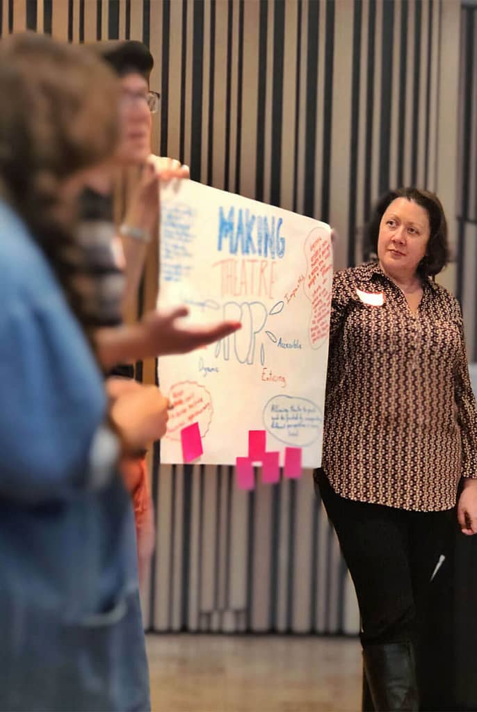 This image shows a group of people holding a poster with colorful writing, with the words ‘Making Theatre Pop’ prominently visible. One person, standing on the right, has light skin and dark hair and is attentively observing the presentation. The poster includes various keywords like ‘Dynamic,’ ‘Engaging,’ and ‘Accessible,’ as well as sticky notes at the bottom, indicating an interactive or brainstorming session focused on theatre creation. The setting appears to be a collaborative workshop or discussion.