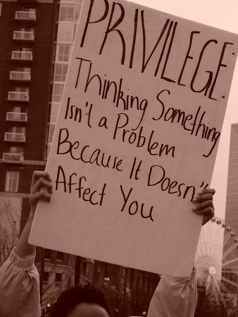 This image shows a person holding up a protest sign that reads: ‘PRIVILEGE: Thinking something isn't a problem because it doesn't affect you.’ The background includes a building and a Ferris wheel, indicating a possible urban setting. The photo is in sepia tone, giving it a vintage or somber feel.