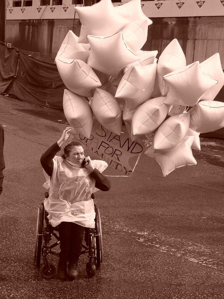 This image shows a person in a wheelchair holding a phone to their ear and a cluster of star-shaped balloons. The person is wearing a rain poncho, and there is a sign attached to the balloons that reads, ‘Stand up for Humanity.’ The background appears to show a city street and some construction barriers.