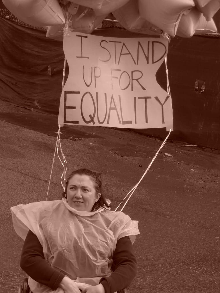 This image depicts a person in a wheelchair wearing a rain poncho and holding balloons with a sign that reads, ‘I stand up for equality.’ The setting appears to be a street, and the person seems to be participating in a demonstration or protest for equality.