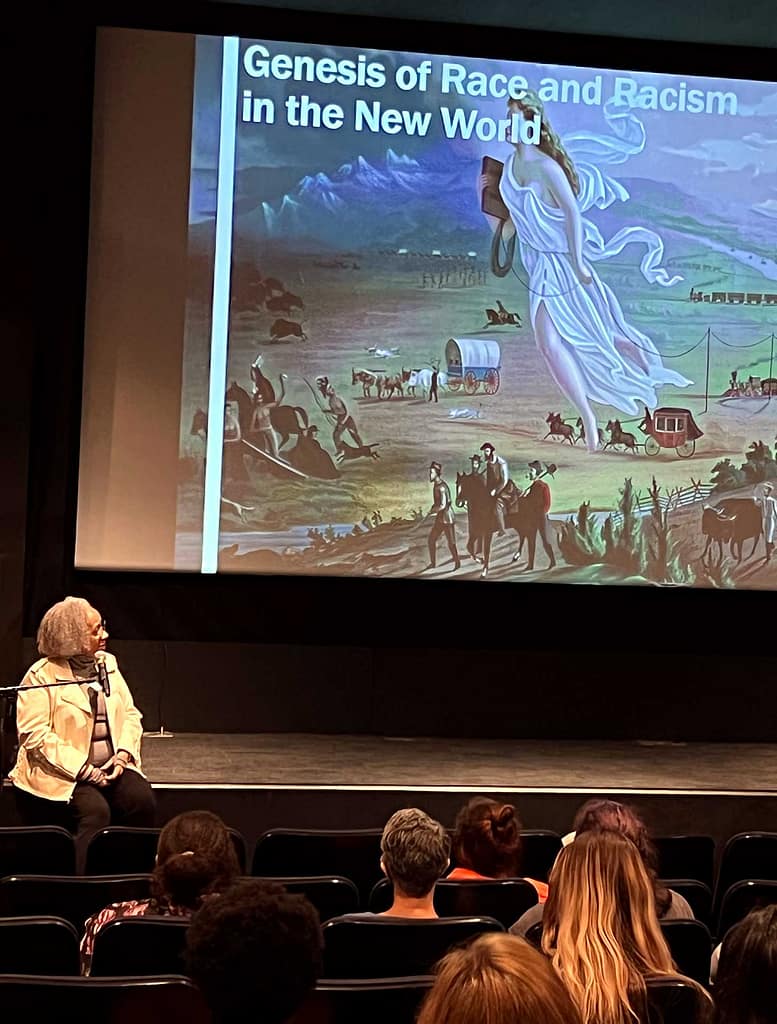 Keryl McCord sitting on a stage in front of an audience, giving a presentation. On the large screen behind her is a slide titled ‘Genesis of Race and Racism in the New World,’ featuring a historic painting of settlers traveling across a landscape with covered wagons and trains. Keryl, who has light gray hair, is seated with a microphone, and the audience is seated in rows of chairs, attentively watching the presentation. The setting appears to be a lecture hall or theater, and the overall atmosphere is academic and educational, focusing on historical themes related to race and racism.