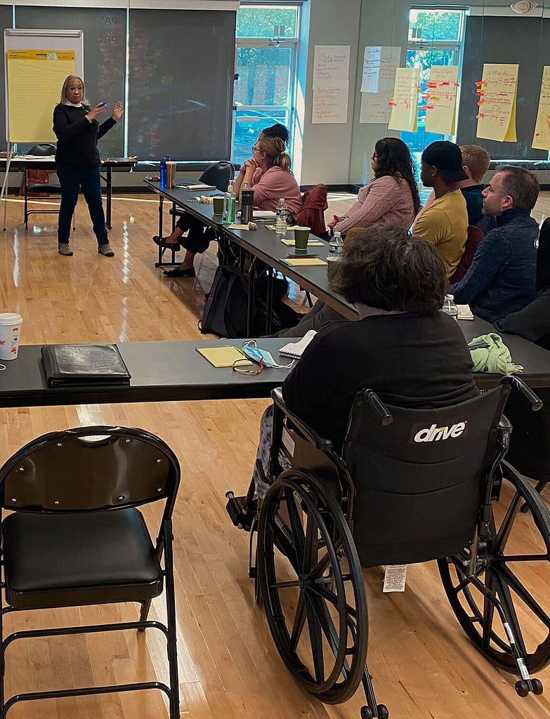 This image shows Keryl McCord, a woman with medium brown skin and short curly gray hair, leading a group discussion in a classroom setting. She stands near a flip chart, gesturing as she speaks, while seated participants listen attentively. The group is seated around long tables, with notepads, cups, and water bottles visible. One participant in the foreground, seated in a wheelchair, faces the front of the room. The atmosphere suggests an interactive workshop or presentation.