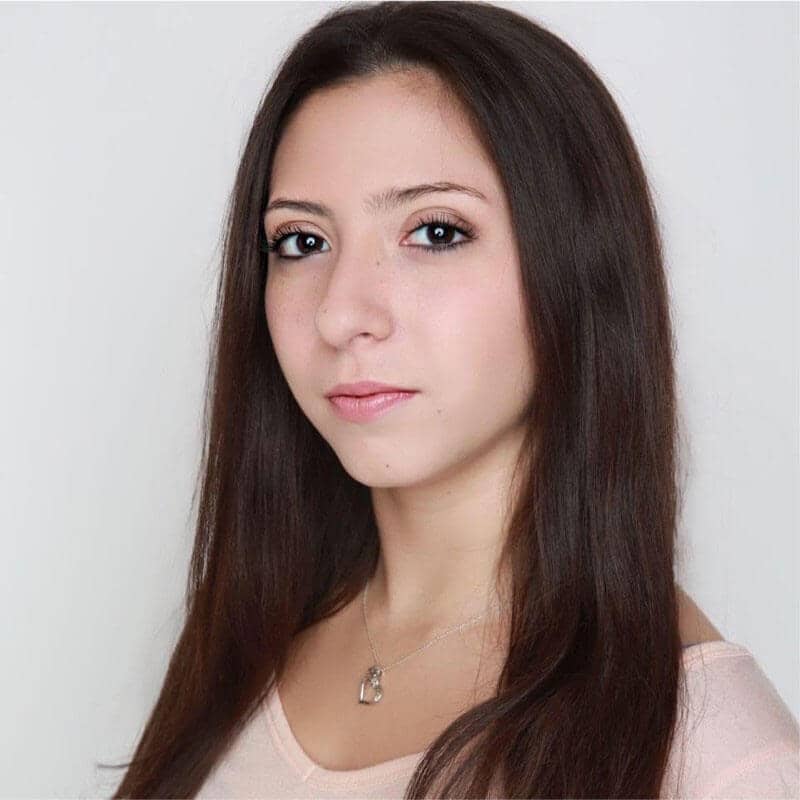 Headshot of Nisa Betancourt, Ta person with long, dark brown hair, wearing minimal makeup and a neutral expression. They are shown from the chest up, have light skin, dark eyes, and are dressed in a light-colored top. A delicate necklace with a small pendant is visible around their neck. The background is plain white, giving the image a clean and professional feel. The individual is looking directly at the camera with a calm and composed demeanor.