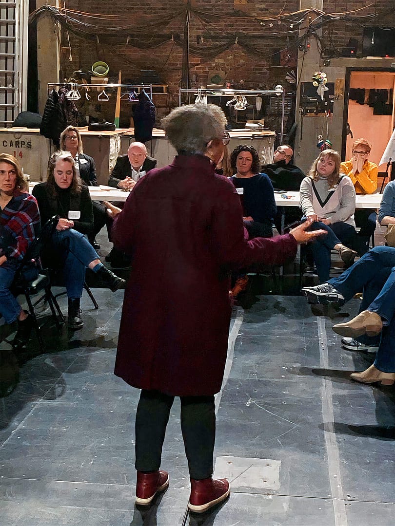 Keryl McCord, a woman with gray hair, wearing a long burgundy coat and red shoes, is standing and speaking to a group of people seated in a semi-circle. The setting is backstage of a theatre with exposed brick walls, ladders, and various equipment in the background. The audience, consisting of individuals with light skin, is attentively listening to her, with some leaning forward and others taking notes. The atmosphere suggests a workshop or an intimate discussion session.
