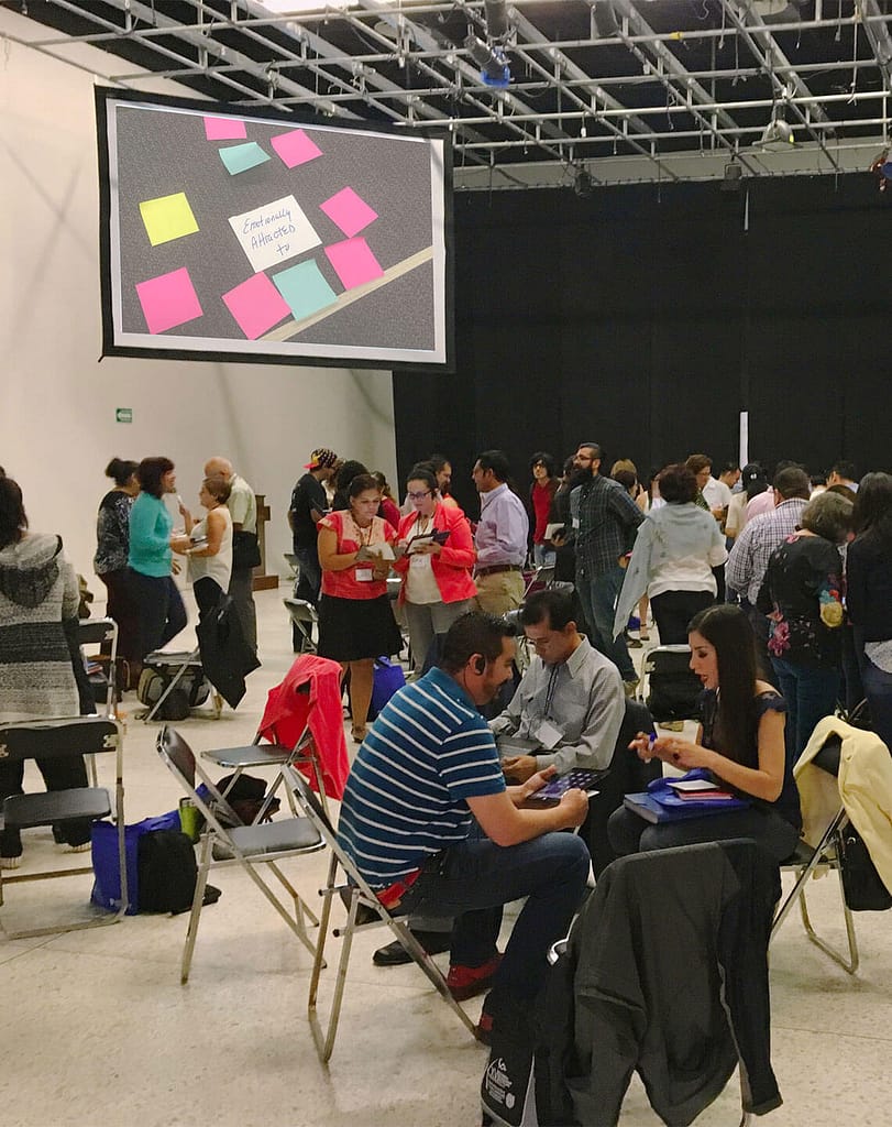 A large group of people with varying skin tones are gathered in a spacious room, engaging in different activities such as discussions and collaborative work. The room has a large projection screen displaying sticky notes with one prominently labeled ‘Emotionally attracted to.’ Several small groups are seated in folding chairs, deeply involved in their tasks, while others stand and converse around the room. The atmosphere suggests a workshop or conference setting focused on teamwork and interaction.