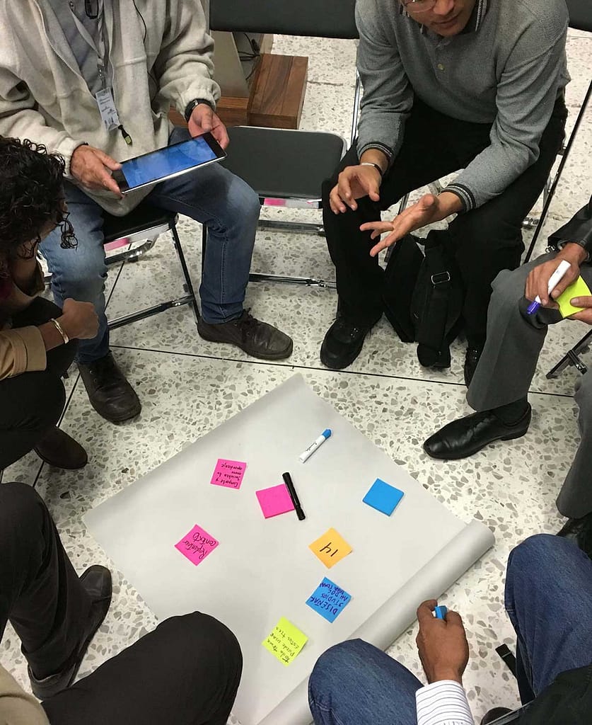 A group of people with varying skin tones are seated in a circle, focusing on a large sheet of paper on the floor in the middle of them. The paper has several sticky notes of different colors, a marker, and a pen on it. One person is holding a tablet, while another is gesturing with their hands, indicating an active discussion or brainstorming session. The setting appears informal, with participants possibly collaborating on ideas or planning.