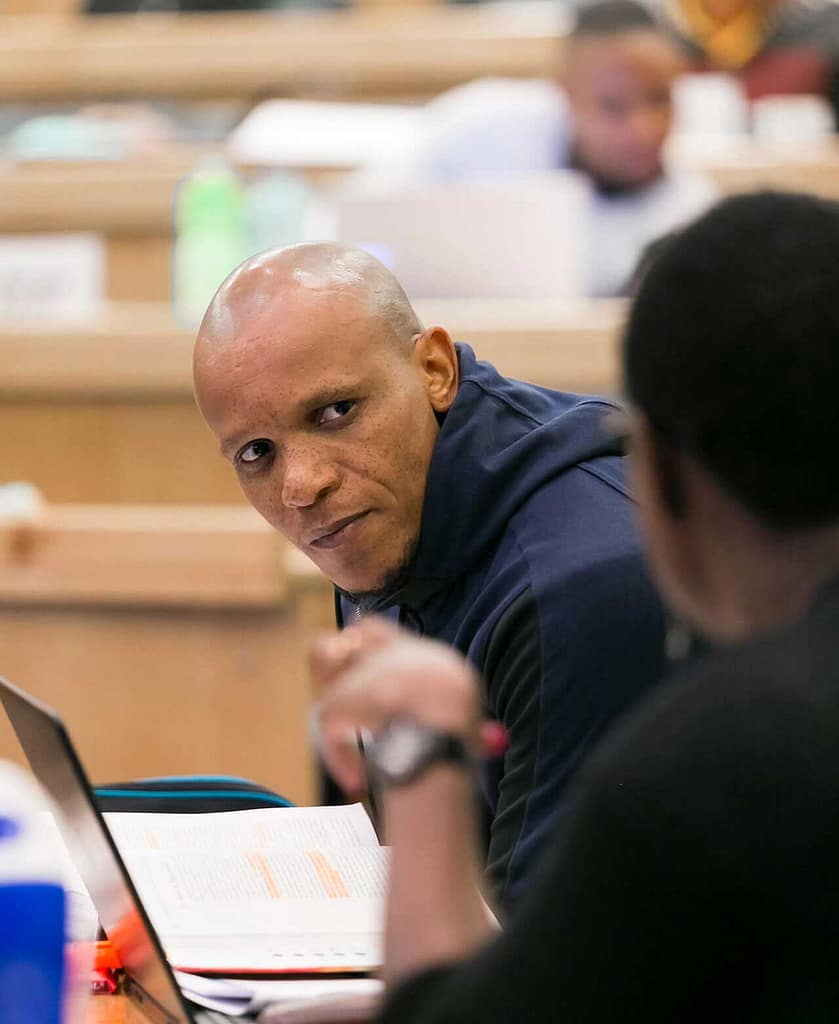 A man with medium brown skin and a shaved head, wearing a dark hoodie, seated in what appears to be a classroom or lecture hall. They are engaged in conversation with another person, whose back is facing the camera. A laptop and open book with highlighted text are visible on the table in front of them. The background shows other people working.