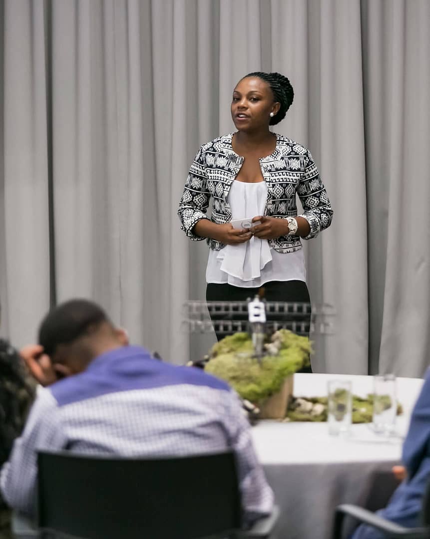 This image shows a woman with medium brown skin and braided hair standing in front of a group, leading a presentation. She is wearing a black and white patterned jacket over a white blouse and appears to be speaking, holding a small note card in her hands. The setting looks like a formal event or conference with tables in the foreground and people seated and listening. The backdrop is a curtain, and the table in front of her has a decorative plant centerpiece.