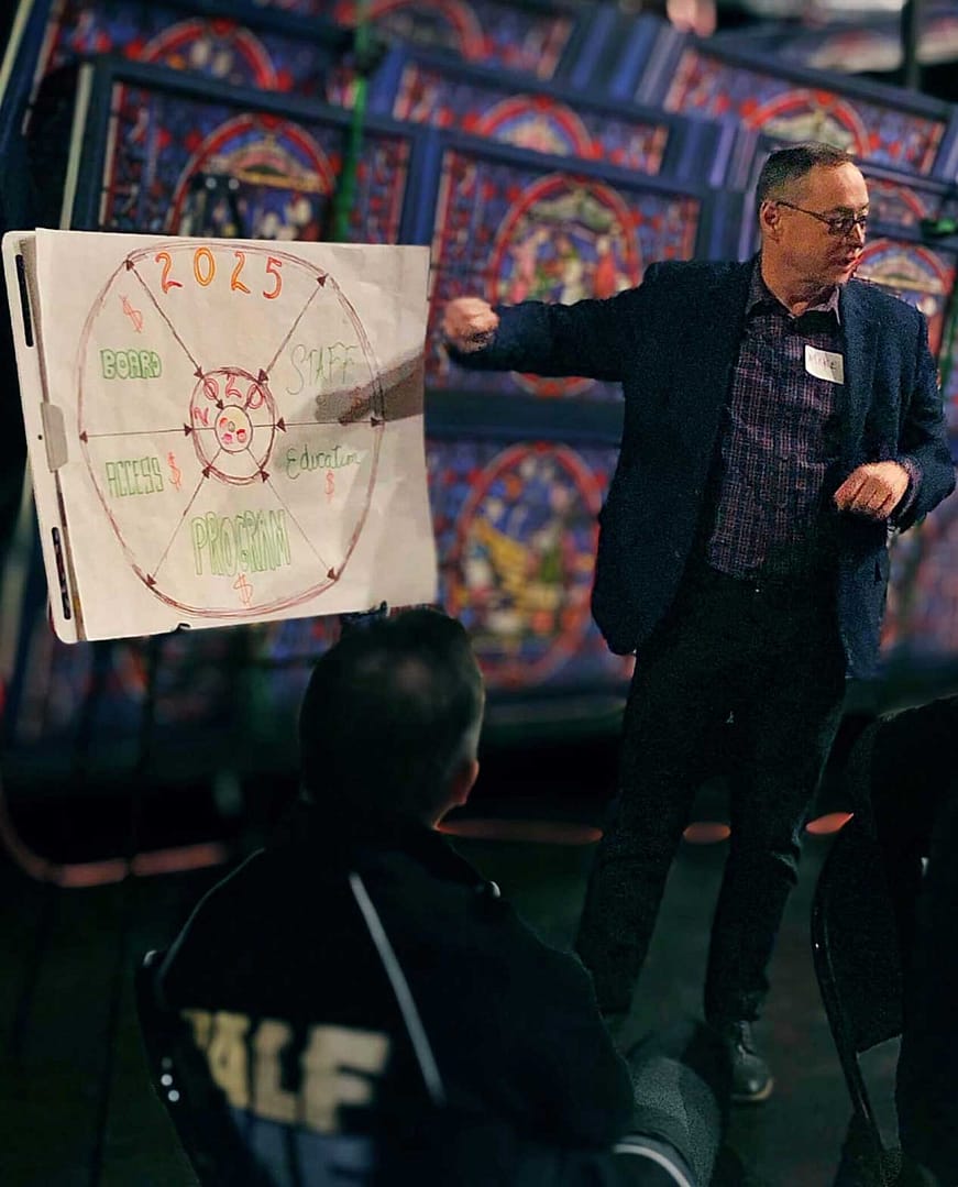 The image shows a man in a dark blazer giving a presentation to a seated audience. He is pointing to a large flip chart that has a circular diagram drawn on it with ‘2025’ at the top. The diagram includes sections labeled ‘BOARD,’ ‘PROGRAM,’ ‘STAFF,’ ‘ASSETS,’ and ‘Education.’ Behind him, a colorful and detailed backdrop, resembling stained glass patterns, adds vibrancy to the setting. The overall scene suggests a collaborative discussion about future plans or strategy development.