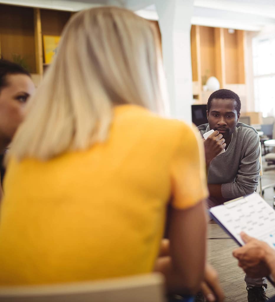 A group of people are engaged in a discussion seated in a circle, with the focus on a dark-skinned man, listening intently. He is leaning forward with his hand on his chin, appearing thoughtful. In the foreground, a light-skinned woman with blonde hair, wearing a yellow top, has her back to the camera. Another person, partially visible, holds a clipboard with notes. The setting is a modern office or meeting room, with shelves and windows in the background, creating a professional and collaborative atmosphere.