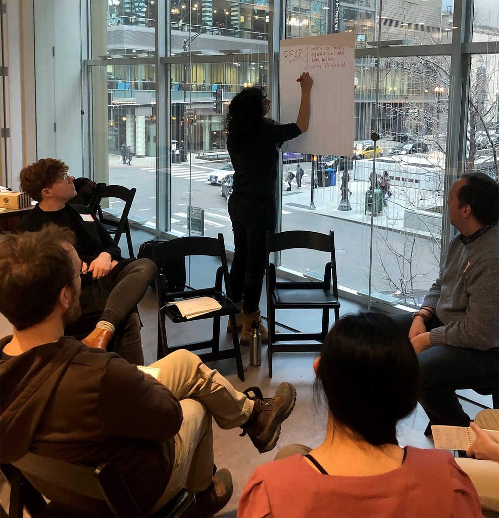 A group of people seated in a semi-circle in a modern space with large glass windows overlooking a city street. A woman dressed in black is standing and writing on a large sheet of paper taped to the window. The paper has the word “FEAR” written at the top, followed by a list of points in red and black marker. The group appears to be engaged in a discussion, possibly part of a workshop or brainstorming session, with some members taking notes while others listen attentively.